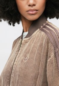 Καφέ ριγωτό μπουφάν μπόμερ από κορδούρα με κλείσιμο με φερμουάρ και αντίθετες καφέ ρίγες στα μανίκια. Διαθέτει κεντημένο λογότυπο στο στήθος.