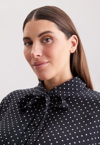 Zwarte blouse met witte stippen, met een geknoopte strik halslijn, een aansluitende kraag en een gladde stoftextuur.