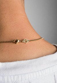 Brățară din lanț de aur cu un pandantiv în formă de floare de lotus, având un lanț texturat și un închizător de tip homar, văzută pe o piele deschisă, purtată cu o cămașă albă.