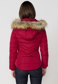 Raudonas paminkštintas striukė su kailiniu gaubtu, pasižyminti įstrižomis siūlėmis ir elastingais rankovės galais. Minkšta tekstūra ir prigludusi forma juosmenyje.