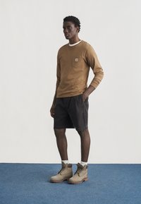 Άνδρας που στέκεται σε μπλε χαλί, φοράει καφέ μακρυμάνικο πουκάμισο, μαύρα σορτς, λευκές κάλτσες και μπεζ μπότες με κορδόνια, μπροστά σε απλό λευκό φόντο.