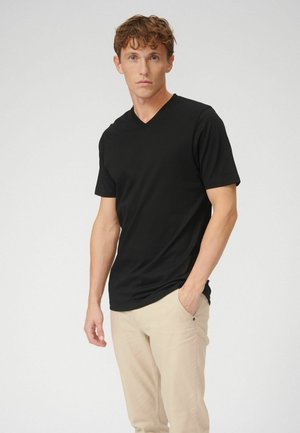 Muž s kučeravými vlasmi, oblečený v čiernom tričku s výstrihom do V a béžových nohaviciach, stojaci s jednou rukou v vrecku na jednoduchom pozadí.