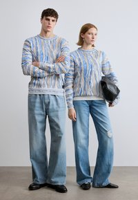 Πλεκτά πουλόβερ με κυματιστό μπλε και μπεζ μοτίβο, συνδυασμένα με ανοιχτό μπλε τζιν παντελόνια φαρδιάς γραμμής. Τα μοντέλα φορούν μαύρα παπούτσια και μαύρη τσάντα.