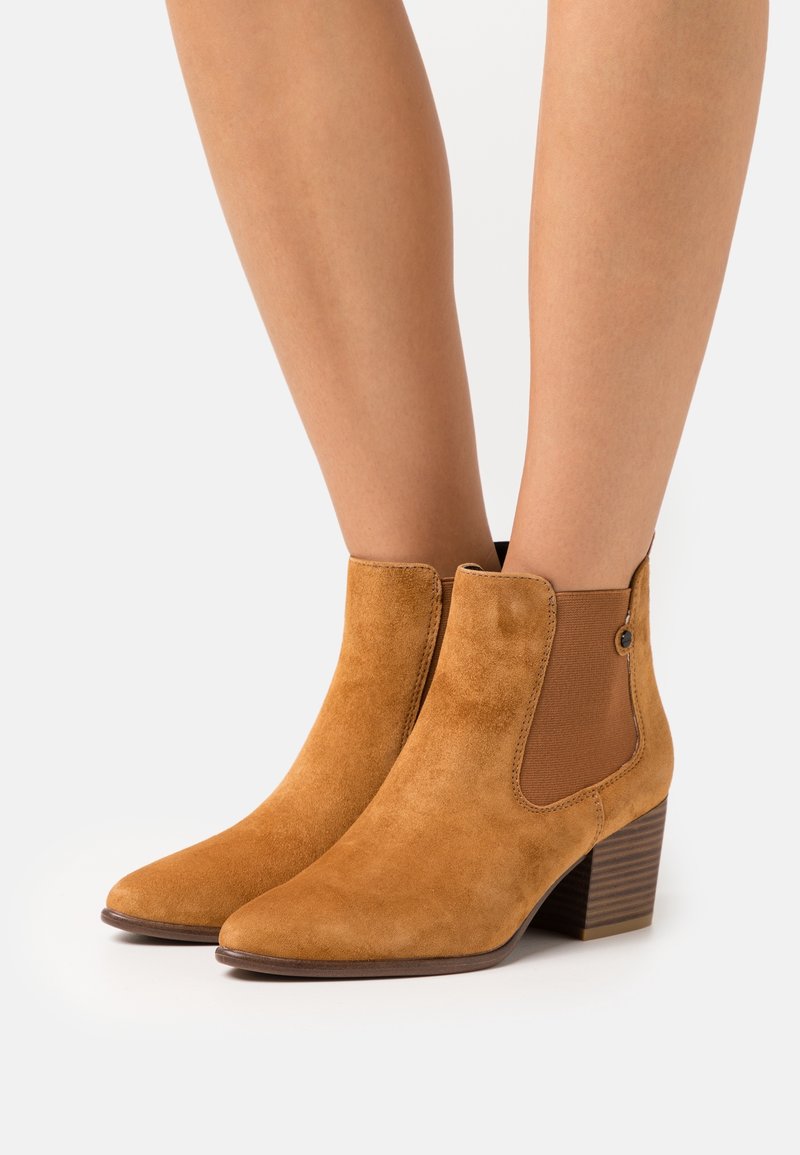 Personne portant des bottines en daim couleur tabac avec talons blocs sur fond gris clair.