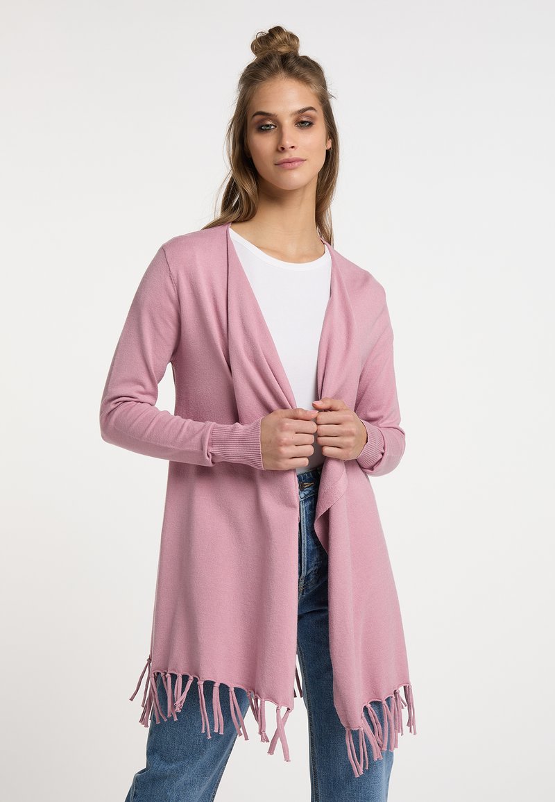 Cardigan rosa chiaro realizzato in tessuto morbido, con un davanti drappeggiato e dettagli a frange sull'orlo. Abbinato a una maglietta bianca e jeans blu.