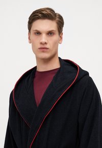 Svart frotté morgenkåpe med hette, med røde detaljer langs kragen og kantene. Båret over en maroon skjorte, med teksturert stoff synlig.
