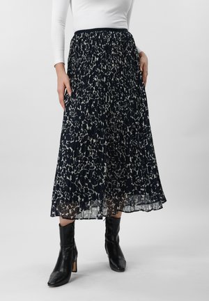 Femme portant une jupe midi plissée à motifs noirs et blancs, un haut blanc à manches longues et des bottines à talons noires, sur un fond uni.