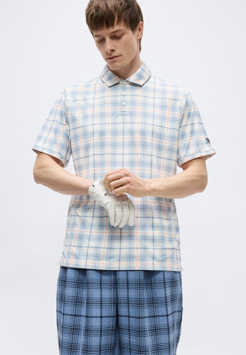 Joven que lleva una camisa de manga corta de cuadros claros y pantalones de cuadros azules, poniéndose un guante de golf blanco en la mano izquierda.