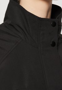 Närbild av en person som bär en svart jacka med hög krage och två svarta tryckknappar fästa nära nacken.