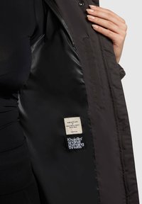 Dark brown coat interior, featuring a smooth texture, a care label, and a brand tag with white text on a solid background.