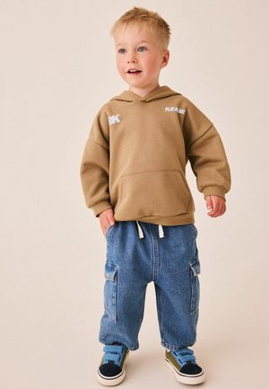 Jeune garçon portant un sweat à capuche marron, un pantalon cargo en denim bleu ample et des baskets bleues et vertes, debout et regardant légèrement vers la droite.