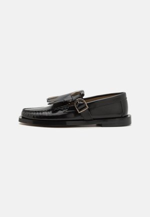 Sapato mocassim de couro preto com um acabamento brilhante, apresentando franjas decorativas e detalhes de fivela. Sola de perfil baixo com aderência mínima.