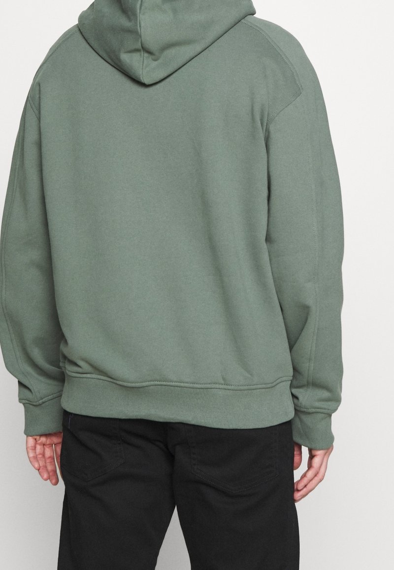 Oliivinvihreä hupullinen collegepaita, jossa on resorit hihansuissa ja helmassa, saumaton selkäosa ja pehmeä, teksturoitu kangas. Yhdistetty mustiin housuihin.