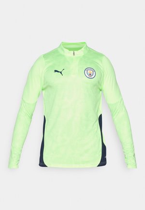 Pitkähiha pullover fluoresoivasta vihreästä kankaasta, jossa on neljännesvetoketju, tummansiniset sivupaneelit ja Manchester City -logo rinnassa.