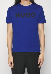 Modrá bavlněná trička s výrazným černým nápisem "HUGO" na přední straně, krátké rukávy a kulatý výstřih.