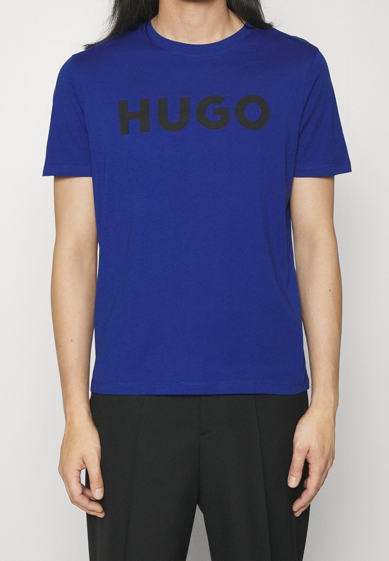 Modrá bavlněná trička s výrazným černým nápisem "HUGO" na přední straně, krátké rukávy a kulatý výstřih.