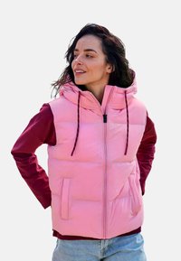 Rosa gesteppte Weste mit Kapuze, vorne mit Reißverschluss, zwei Seitentaschen und dunkelroten langen Ärmeln. Glatte Textur mit kontrastierenden Farben.