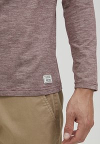 Langærmet burgunder T-shirt med et struktureret mønster og et hvidt mærke på kanten. Båret med beige bukser, der fremhæver en afslappet pasform.