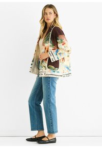 Quilted jacket in light cream, featuring colorful artistic patterns, brown accents, and black label details. Paired with blue jeans and black flats.