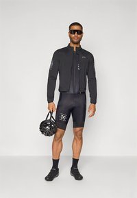 Svart sykkeljakke med tettsittende design, kombinert med svarte shorts med logodetaljer; modellen holder en svart hjelm.