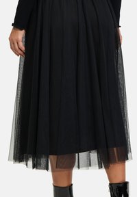 Gonna di tulle nera con pieghe, lunghezza alla caviglia, caratterizzata da una trama morbida e un design a strati. Indossata con un top nero aderente e stivali neri lucidi.