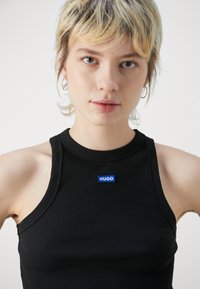Black ribbed tank top with a high neckline and blue "HUGO" logo. Features sleeveless design and textured fabric.