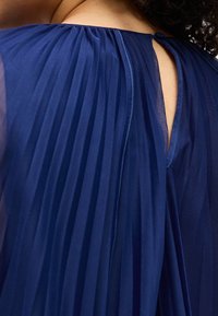 Σκούρο μπλε πλισέ ύφασμα που είναι ριγμένο πάνω στην πλάτη ενός ατόμου, με μια μικρή τρύπα στο σημείο του λαιμού και κατσαρά μαλλιά στην κορυφή.