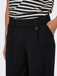 Pantalon noir en tissu lisse et texturé, coupe ajustée, avec boutons sur la ceinture et poches latérales.