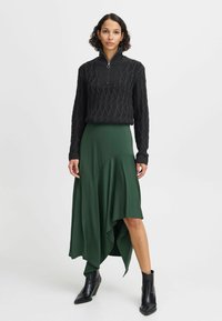 Svart stickad tröja med halvdragkedja, kombinerad med en grön asymmetrisk omslagskjol. Modell bär svarta ankelstövlar.