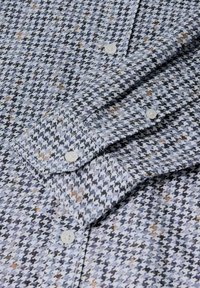 Camicia con motivo houndstooth blu chiaro e marrone, con bottoni bianchi, che mostra in dettaglio la parte frontale e il polsino piegato.