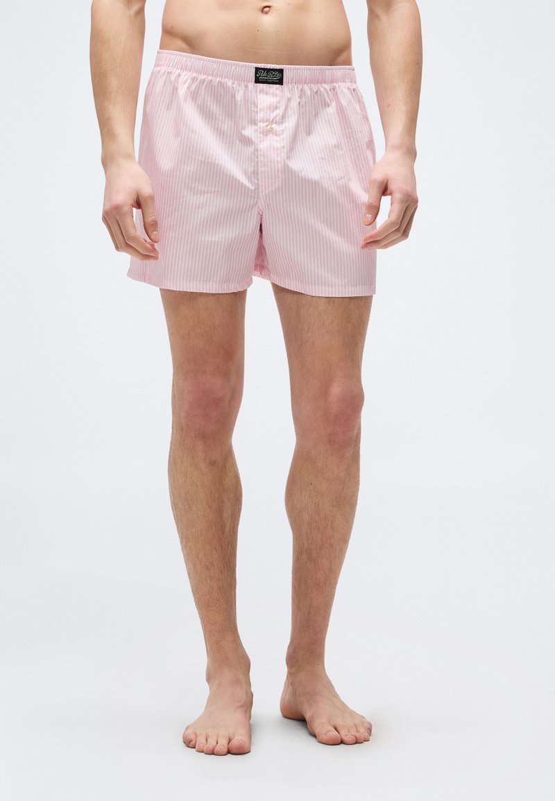 Άνδρας που φοράει ανοιχτό ροζ και λευκά κάθετα ρ ιγέ μποξεράκια, όρθιος με γυμνά πόδια μπροστά σε απλό φωτεινό φόντο.