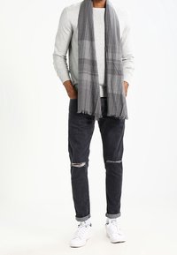 Grau gestreifter Schal mit kariertem Muster, getragen über einem hellgrauen Pullover, kombiniert mit dunklen, zerrissenen Jeans und weißen Sneakern. Lässiges Outfit.