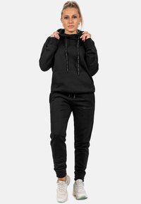 Felpa nera con cappuccio e lacci, abbinata a pantaloni della tuta neri coordinati. Presenta polsini elasticizzati e una vestibilità comoda.
