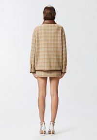 Chaqueta de tweed de cuadros marrón con un cuello de cuero marrón, combinada con una falda de cuadros a juego. Sandalias de tacón blanco completan el conjunto.