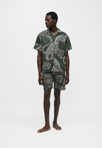 Άνδρας στέκεται ξυπόλητος φορώντας σκούρο πράσινο ταιριαστό κοντομάνικο πουκάμισο και σορτς με λευκό λεοπάρ μοτίβο με κηλίδες.