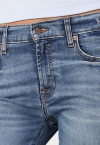 Jeansi din denim albastru închis cu o textură ușor uzată, având accesorii din cupru și cusături portocalii, și un design standard cu cinci buzunare.