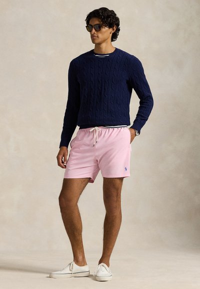 Moški v temno modrem izdelanem puloverju, roza kratkih hlačah z vrvico, belih supergah, sončnih očalih in ročni uri, stoji ob preprostem ozadju.
