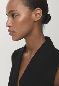 Giovane donna con pelle liscia di colore marrone e capelli raccolti in uno chignon, che indossa un indumento senza maniche di colore nero, rivolta a sinistra su uno sfondo neutro.