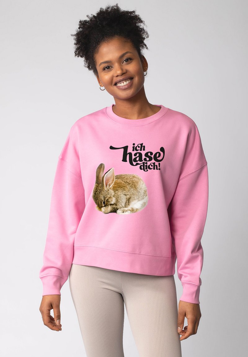 Lächelnde Frau mit lockigem Haar trägt ein pinkes Sweatshirt, auf dem ein brauner Hase und der deutsche Text "ich hase dich!" zu sehen sind, vor einem einfarbigen Hintergrund.