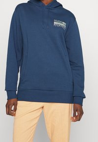 Felpa blu navy con cappuccio e cordino, dotata di tasca a marsupio frontale. Presenta un grafiche del logo "HUGO" stampato sul petto sinistro. Tessuto morbido al tatto.