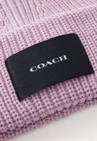 Berretto lavorato a maglia rosa con una trama testurizzata, dotato di un'etichetta rettangolare nera con la scritta bianca "COACH". Materiale morbido e caldo.