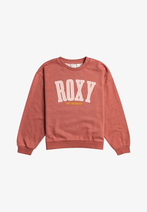 Femme portant un sweat-shirt rose poudré "ROXY Surf University", un pantalon de jogging gris et des baskets blanches avec des accents pastel. Mains dans les poches.