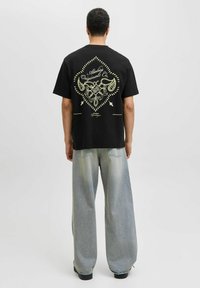 Sort bomulds T-shirt med et gult dekorativt design på ryggen, parret med brede lyseblå jeans og sorte fodtøj.