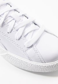 Weißer Sneaker mit glänzendem synthetischem Obermaterial, strukturiertem Gummisohle, flachen weißen Schnürsenkeln und dezenten Nähten an den Seiten.