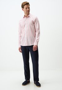 Chemise blanche à rayures verticales rouges, à col bouton, à manches longues. Portée avec un pantalon bleu marine foncé et des chaussures à enfiler bleues marines, dans une posture debout.