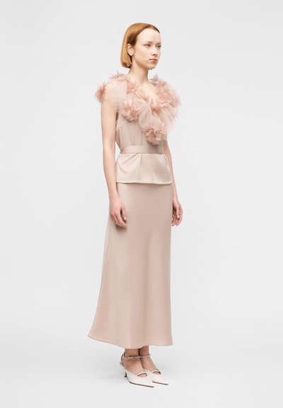 Robe longue ajustée beige avec un corsage structuré et un col transparent à volants. Associée à des mules blanches à talons avec des découpes.