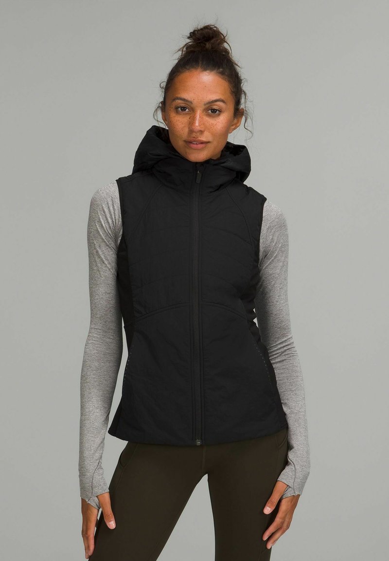 Gilet nero con cappuccio, zip, design trapuntato e tasche laterali, indossato sopra una maglietta a maniche lunghe grigia chiara. Tessuto strutturato con una vestibilità sportiva.