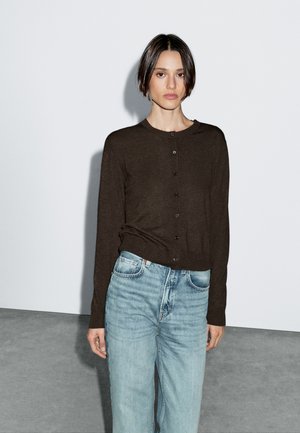 Woman with short dark hair wearing a buttoned dark brown cardigan and light blue high-waisted jeans standing against a plain white wall.