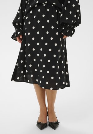 Schwarzer Midirock mit weißen Punkten, kombiniert mit passendem Oberteil und schwarzen spitzen Schuhen, Hände seitlich am Körper.
