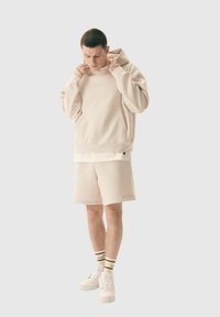 Hoodie oversize beige avec poche avant, associé à un short assorti. Présente un branding subtil et un tissu texturé et confortable. Baskets blanches et chaussettes rayées.